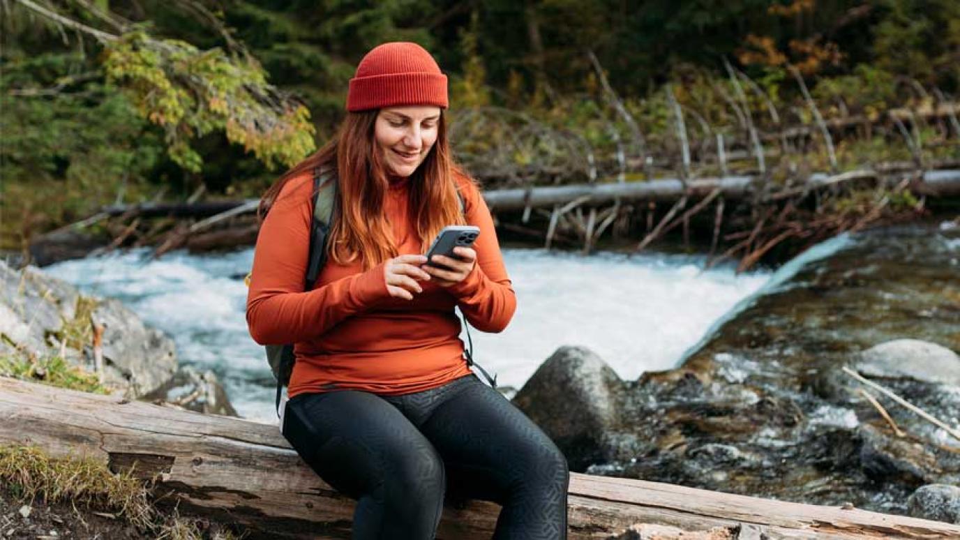 Happy redhead Woman in red hat stopping for a break while hiking to look at map on her phone. A fast flowing river winds behind her.