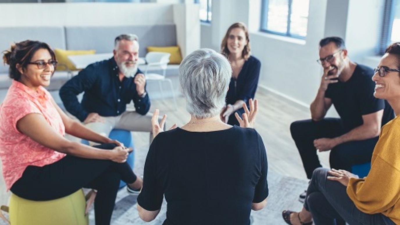 A group of smiling adults gathered around a woman, listening to her.