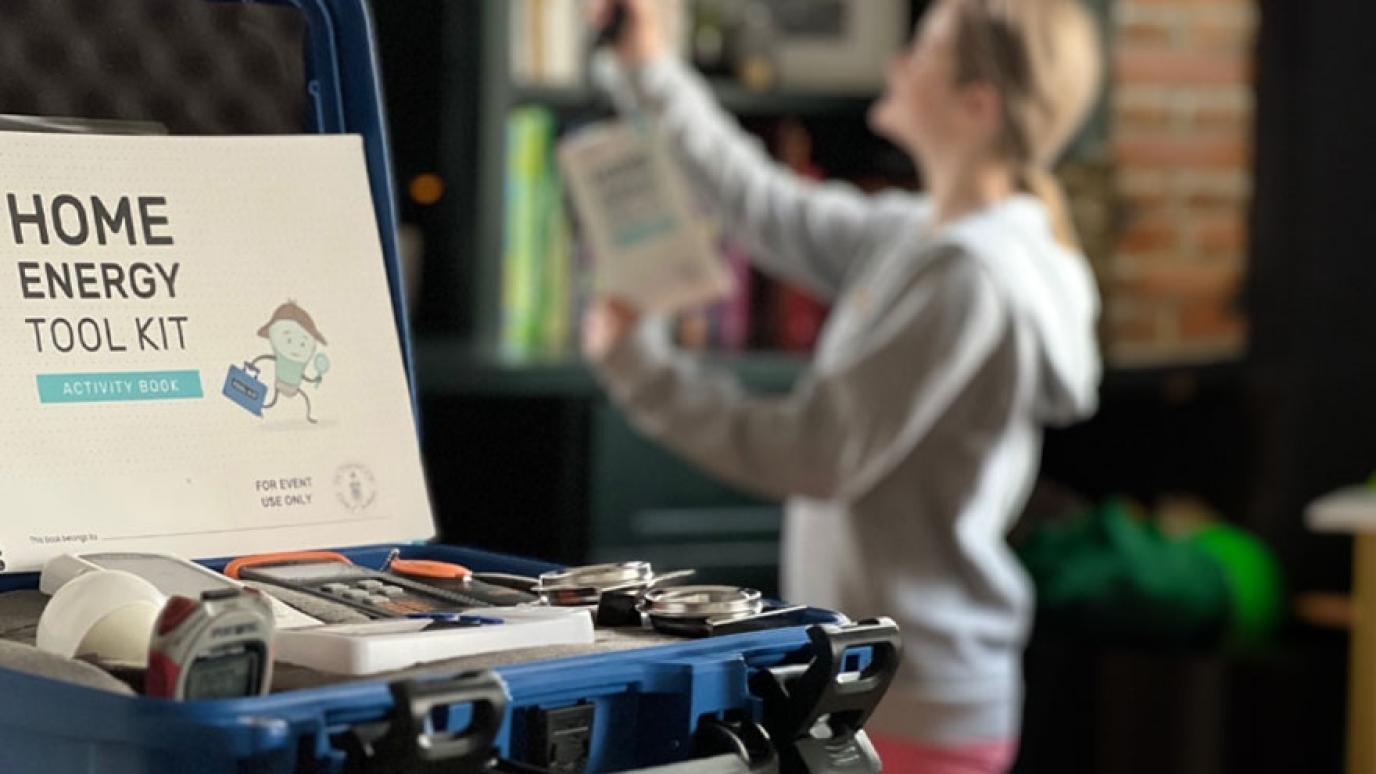 Home Energy Toolkit full of energy tools sits on a table in the foreground. A blurred figure in the background uses one of the tools to check a reading in their home.