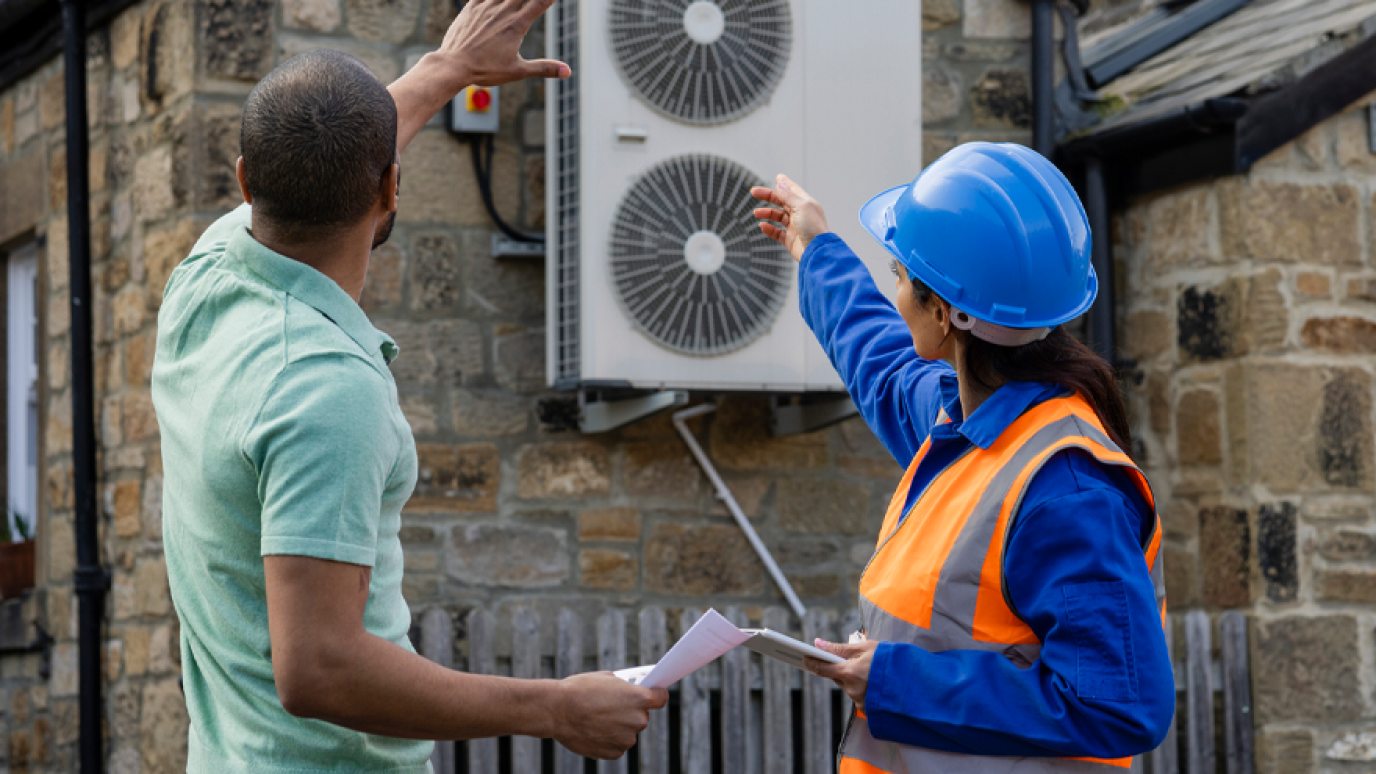 A worker with a hard hat and reflective jacket consults with a homeowner. They are both pointing to a heat pump.