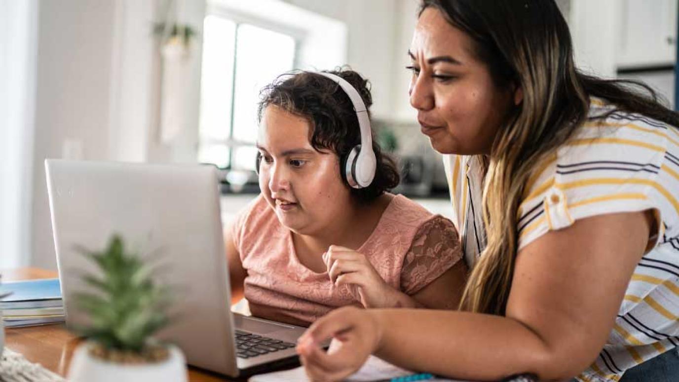 Mother assists daughter with special needs with her work on a laptop. Child is wearing headphones.