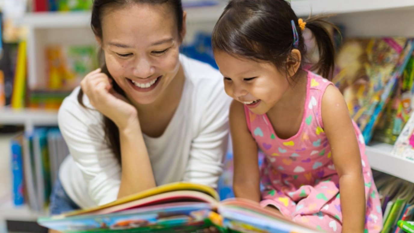 Mother and daughter laughing with each other as they read from a picture book at a local public library.