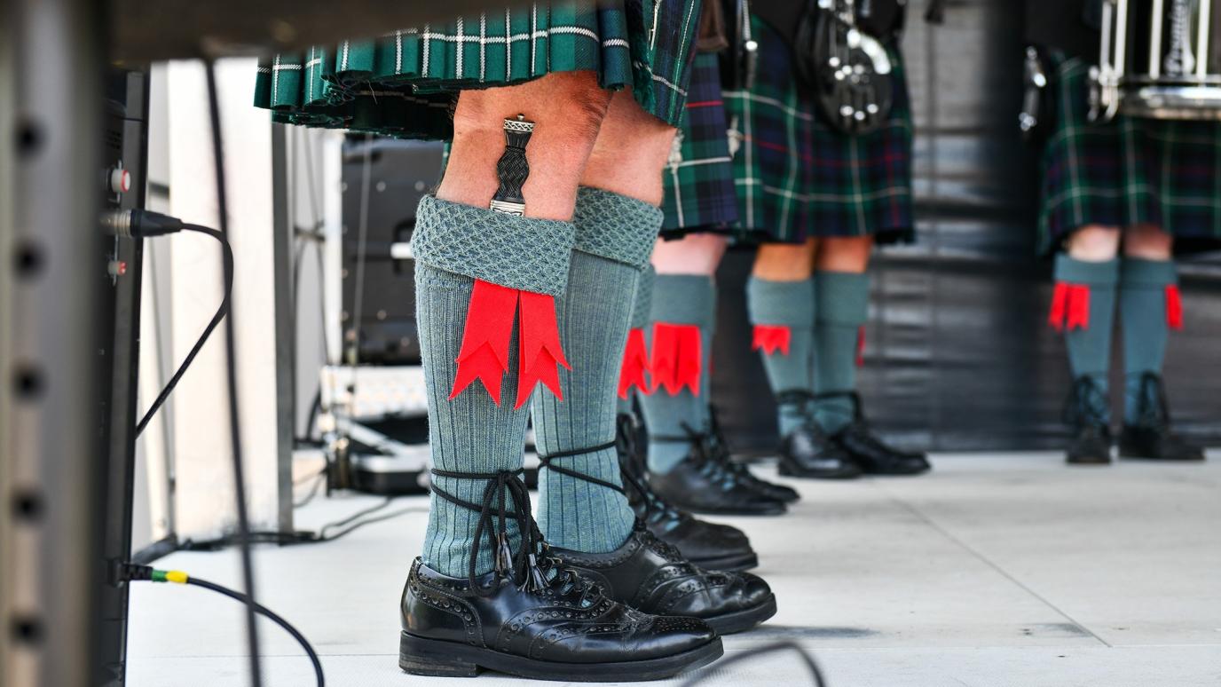 Close-up of performers wearing kilts, green socks, and black shoes with flashes of red ribbon, part of a traditional Scottish pipe and drum band.