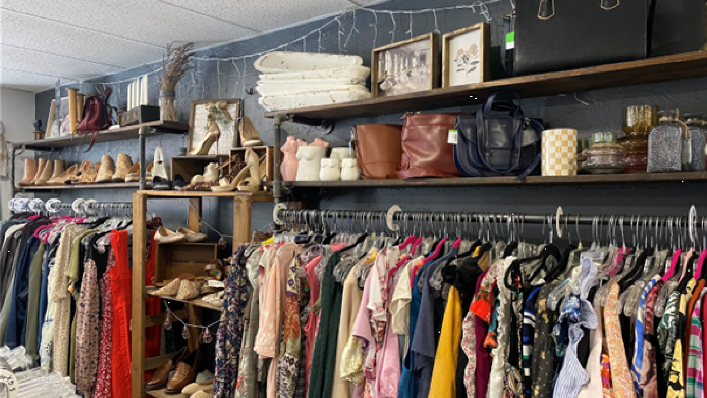 A corner of a thrift store with various clothes hanging on racks. Nearby shelves display assorted items such as shoes, bags, art and much more.