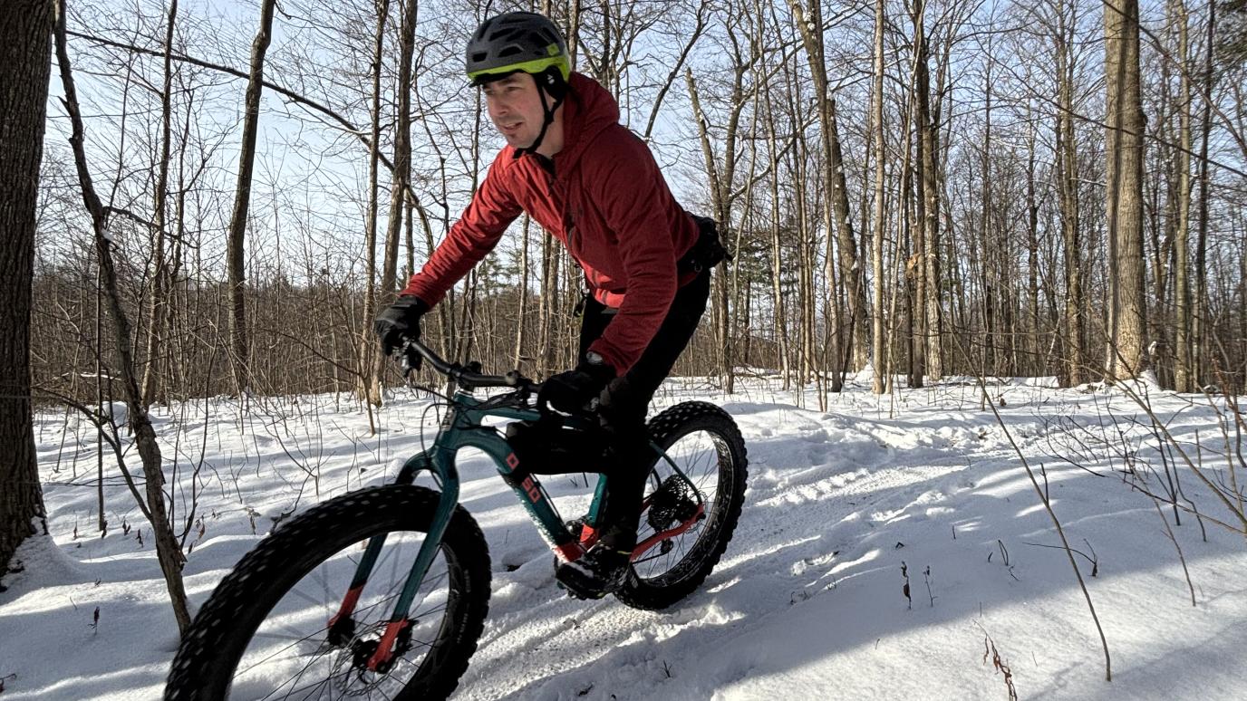 A person wearing a red jacket and helmet rides a fat-tire bike along a narrow snow-covered trail through a forest in winter