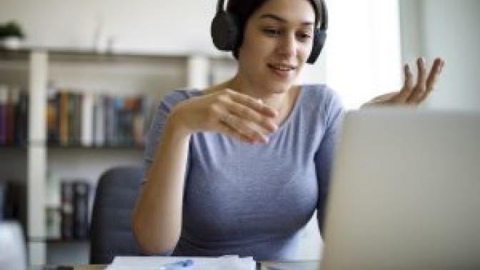 A woman with headphones on looks down toward a laptop on the table in front of her. 