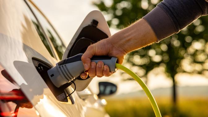 Hand of man inserting a power cord into an electric car for charging ecofriendly vehicle on green landscape