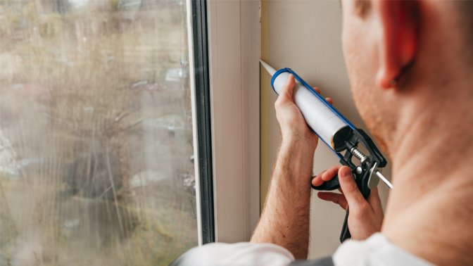 Young man wearing overalls sealing cracks between window and trim using waterproof silicone caulk on the balcony.