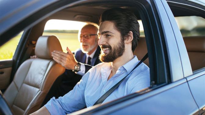 A young man drives a rideshare vehicle through a rural area talking happily with his passenger in the back seat
