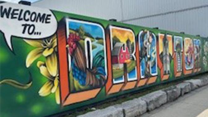 Large, colourful mural on outdoor wall saying 'Welcome to Drayton' surrounded by imagery of flowers and town landmarks.