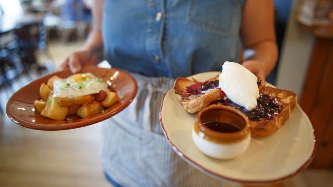 A waitress in a blue uniform serves plates of pancakes, eggs, hashbrowns and bacon to customers at a local diner
