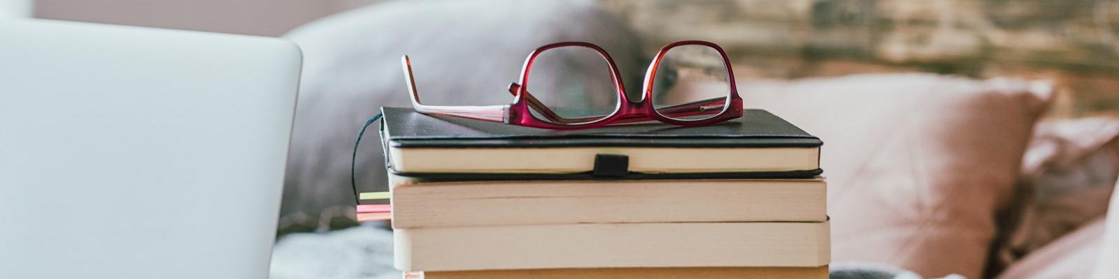 A stack of books with a pair of glasses resting upside down on top.