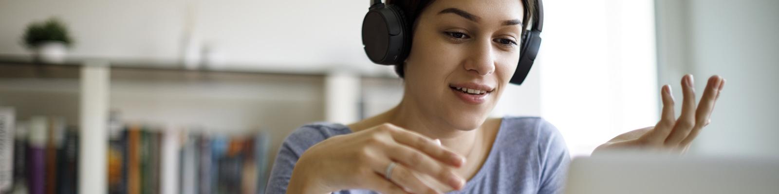 A woman with headphones on looks down toward a laptop on the table in front of her. 