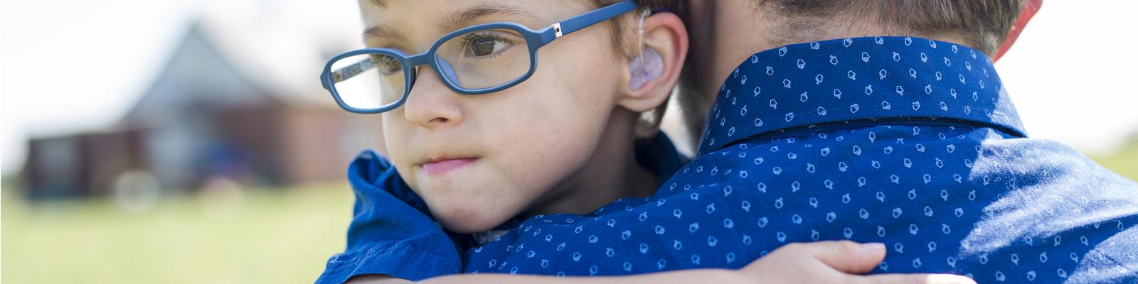 A young boy with glasses and a hearing aid is held by his father, looking behind him over his shoulder.