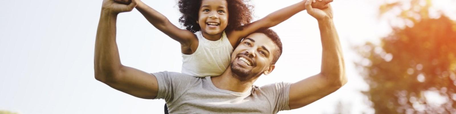A father holds his daughter on his back, holding her hands outstretched. They both smile. 