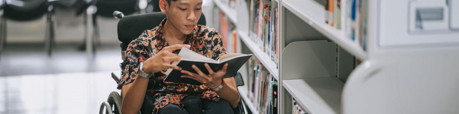 Teenage boy wheelchair reading book in library