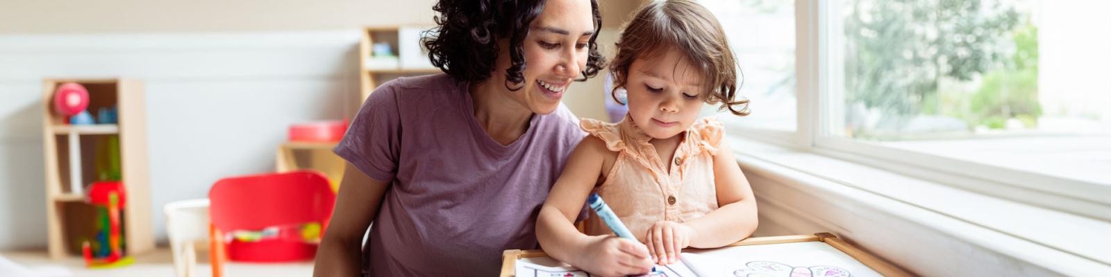 Caregiver smiling over child working on colouring pages in a child care setting