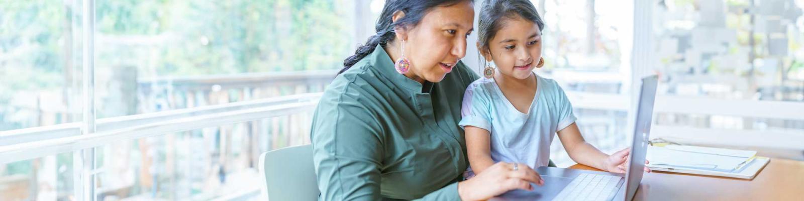 Mom with young daughter on her lap reviewing something on a laptop at a kitchen table