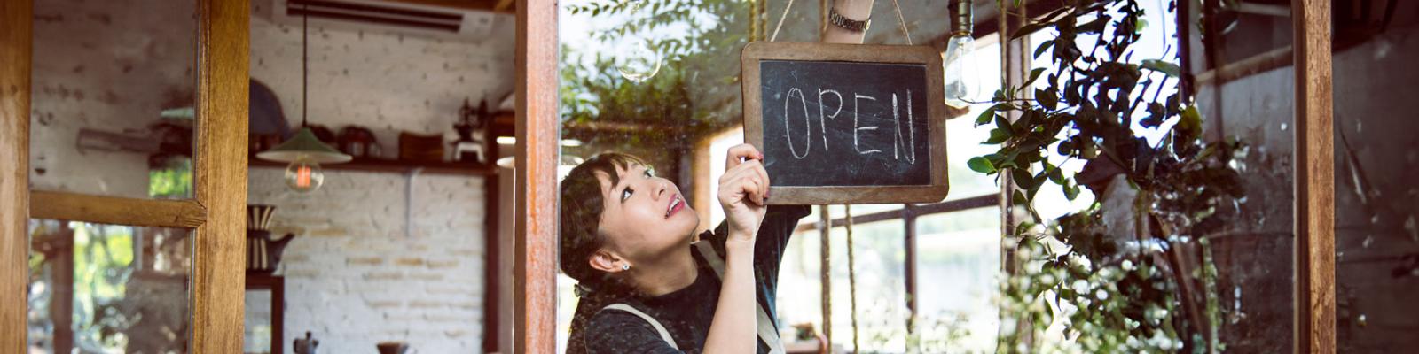 young woman hanging an open sign in the window of her business