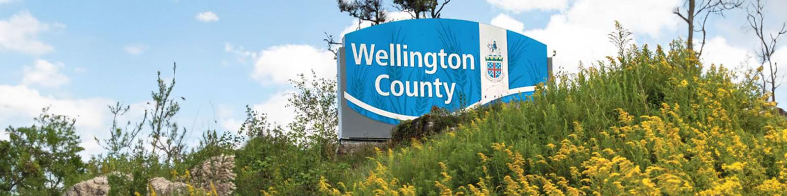 Wellington County blue and white Welcome sign on a grassy slope next to a roadway
