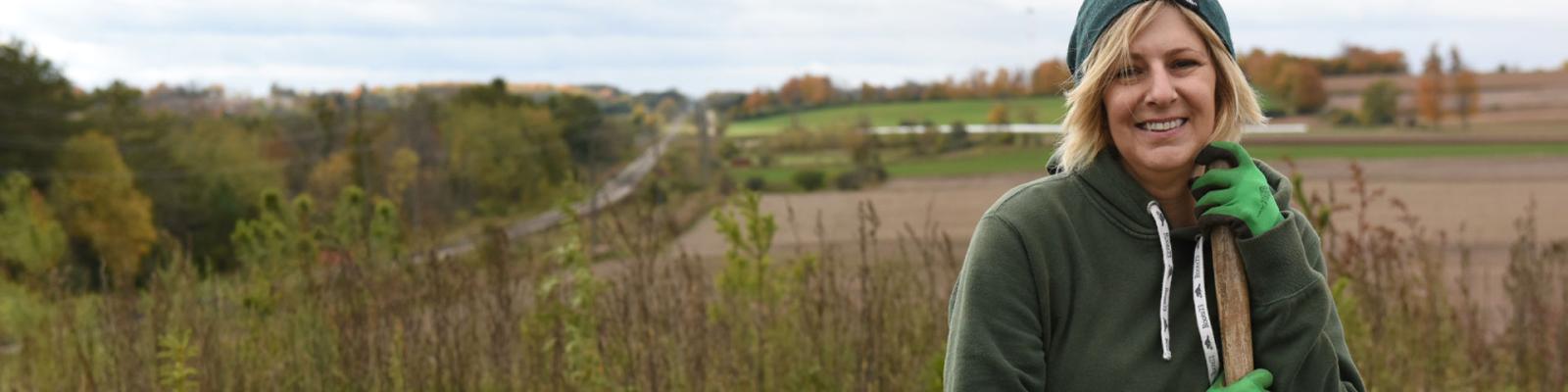 Close up crop of woman smiling, leaning on handle of her shovel while tree planting. Rolling rural fields in the background