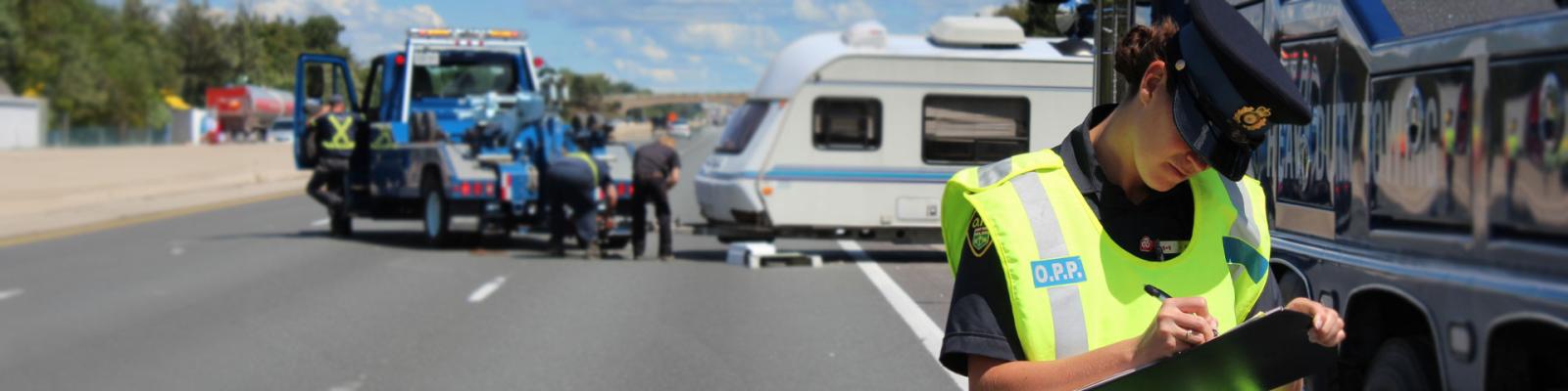 OPP officer in bright yellow safety vest writing report on scene of an accident on a divided highway. Accident is blurred in background.