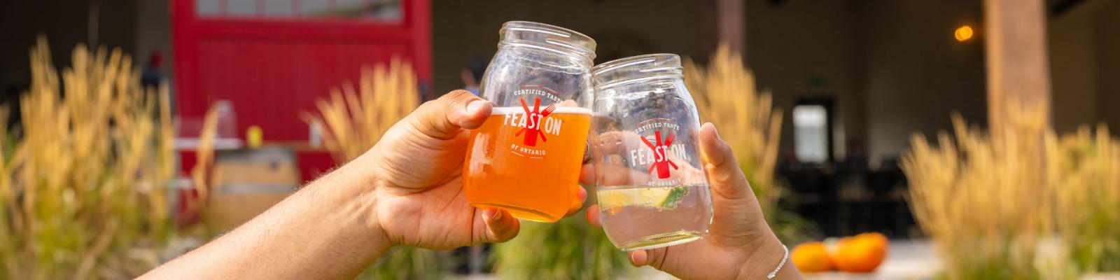 Two hands doing a cheers with mason glasses farm scene in background