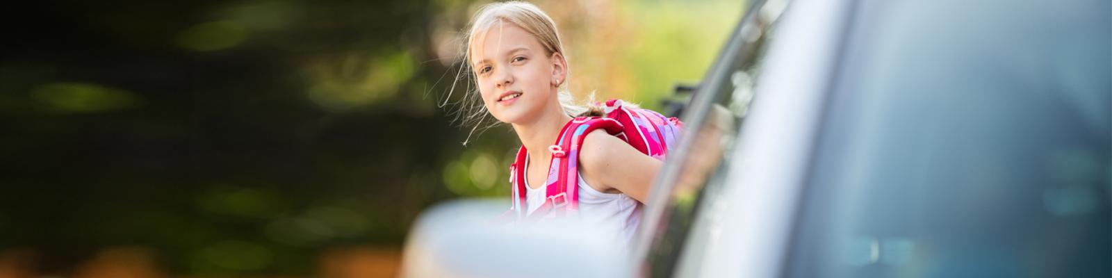 A young girl wearing a backpack crosses the street in front of a stopped car