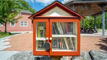 Photograph of an orange little library filled with dozens of books standing in front of a pavilion with picnic tables. 
