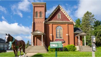 Photograph of a brown brick building with a statue of a brown horse stands in the front walkway, next to a green historical plaque. 