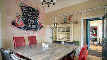 Photo of the interior of a cafe with a large black top hat mural painted on the wall and filled with text. A table with red chairs sits in the middle of the room under a chandelier. 