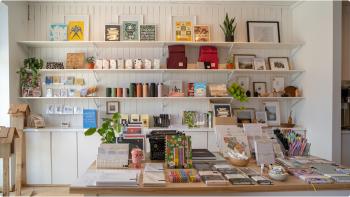 Interior of a store with white paneled walls and shelves, full of small home items, cards, stationary, artwork, and more. 