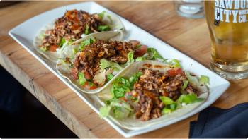 Three jerk chicken tortilla tacos sit on a white plate next to a light beer.