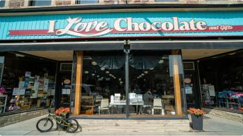 The outside of a storefront with a large blue, white and red sign that reads “I Love Chocolate and Gifts” above a double window, with a bike outside of it. 