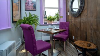Small table with large purple chairs set with two wine glasses in a room with purple window frames, plants, and a wooden decorative wall. 