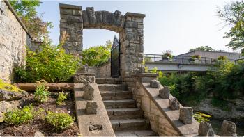 Stone steps leading up to a stone archway with a large black gate, next to a bridge and flower gardens. 
