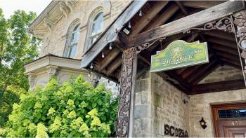 Outdoor photograph of a stone house surrounded by greenery with a covered front porch and a green hanging sign that says n “The Breadalbane Inn.”