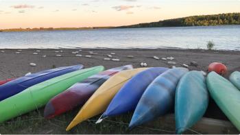 Nine colourful canoes sit on a sandy beach in front of a lake with trees and a sunset in the distance. 