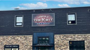 A sign with 'Chompin at The Bit Bar and Grill' above a set of blue restaurant doors, with cloudy blue skies above. 