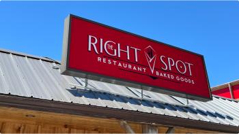 Image of a red sign that says "The Right Spot Restaurant and Baked Goods" sitting on top of a grey roof above a wooden storefront with blue skies in the background.