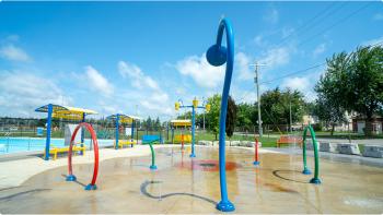 Image of an empty splash pad with an assortment of red, blue, and yellow sprinklers spraying water under a bright blue sky. Text reads: On a hot summer day I like to - Splash! Nothing makes my heart happier than a visit to the Mouth Forest or Arthur community splashpads for a quick cool down. Follow it up with some deep-fried pickles and a submarine sandwich from Erni's Place and I'm in heaven.