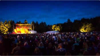 Dark blue sky above a large crowd of people watching a band perform on a stage with bright yellow and orange lights.