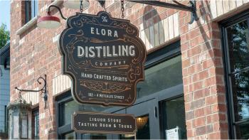 Close up of a hanging storefront sign reading “Elora Distilling Company” in front of a red brick building. 
