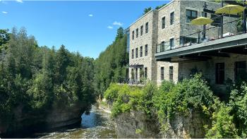 View of the Grand River flowing by a long path of green trees, next to the Elora Mill Spa’s stone building and outdoor deck and patio.