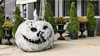 A large paper sculpture of a pumpkin adorned with lights sits in front of a patio with white tables, green trees, and black planters. 