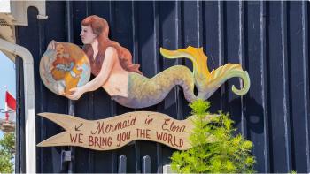 Sign of a mermaid holding a globe in her hands with the words “Mermaid in Elora, We bring you the world” mounted to the side of building with navy blue siding. 