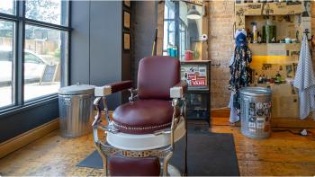 Image of a burgundy barber’s chair in the middle of a room with yellow brick walls, hardwood floors, wall mounted shelving covered with old newspaper clippings, and a large bright window. 