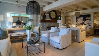 Image of a designed living space featuring a stone fireplace surrounded by stylish white furniture, a glass coffee table with white flowers, and a large pendant light hanging from the ceiling. 