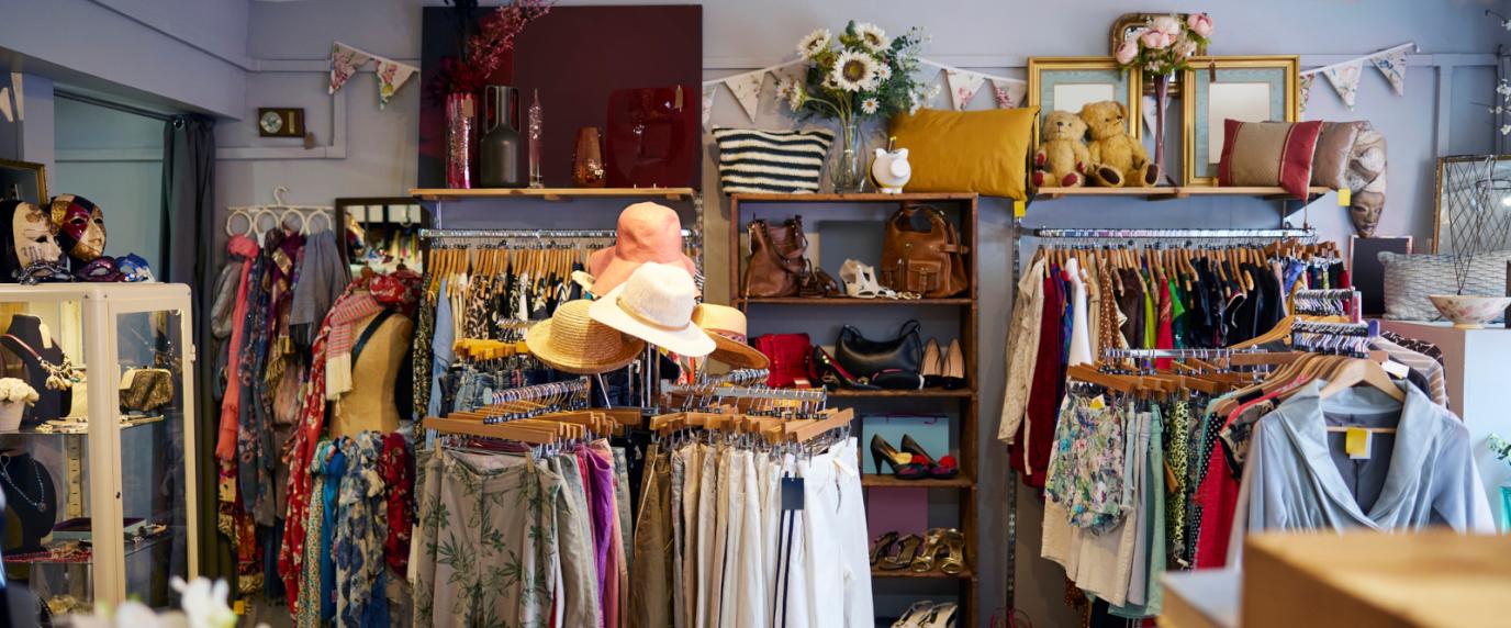 A cozy and colorful thrift store interior with neatly arranged racks of second-hand clothing, hats, handbags, and shoes. The store features decorative items such as pillows, flowers, framed mirrors, and plush teddy bears on upper shelves, creating a welcoming and eclectic atmosphere. A display case on the left showcases jewelry and accessories.