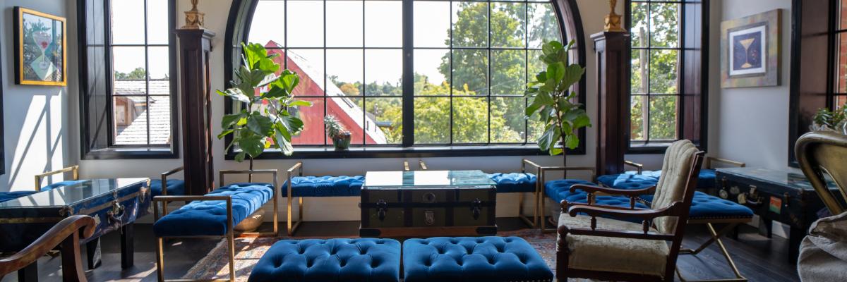 A bright, elegant lounge with arched windows, rich blue velvet seating, and vintage-modern decor. Lush plants and artwork add charm to this stylish, inviting space.